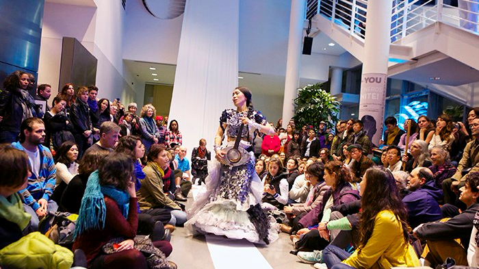 A performer travels down a narrow pathway between audience members seated on the right and left. They wear an elaborate dress with ruffled skirts and seem to be playing a stringed instrument.
